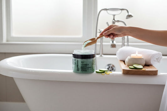 A jar of cucumber splash bath with cucumber peel and a black lid, accompanied by sliced cucumbers and a few drops of a yellow substance.