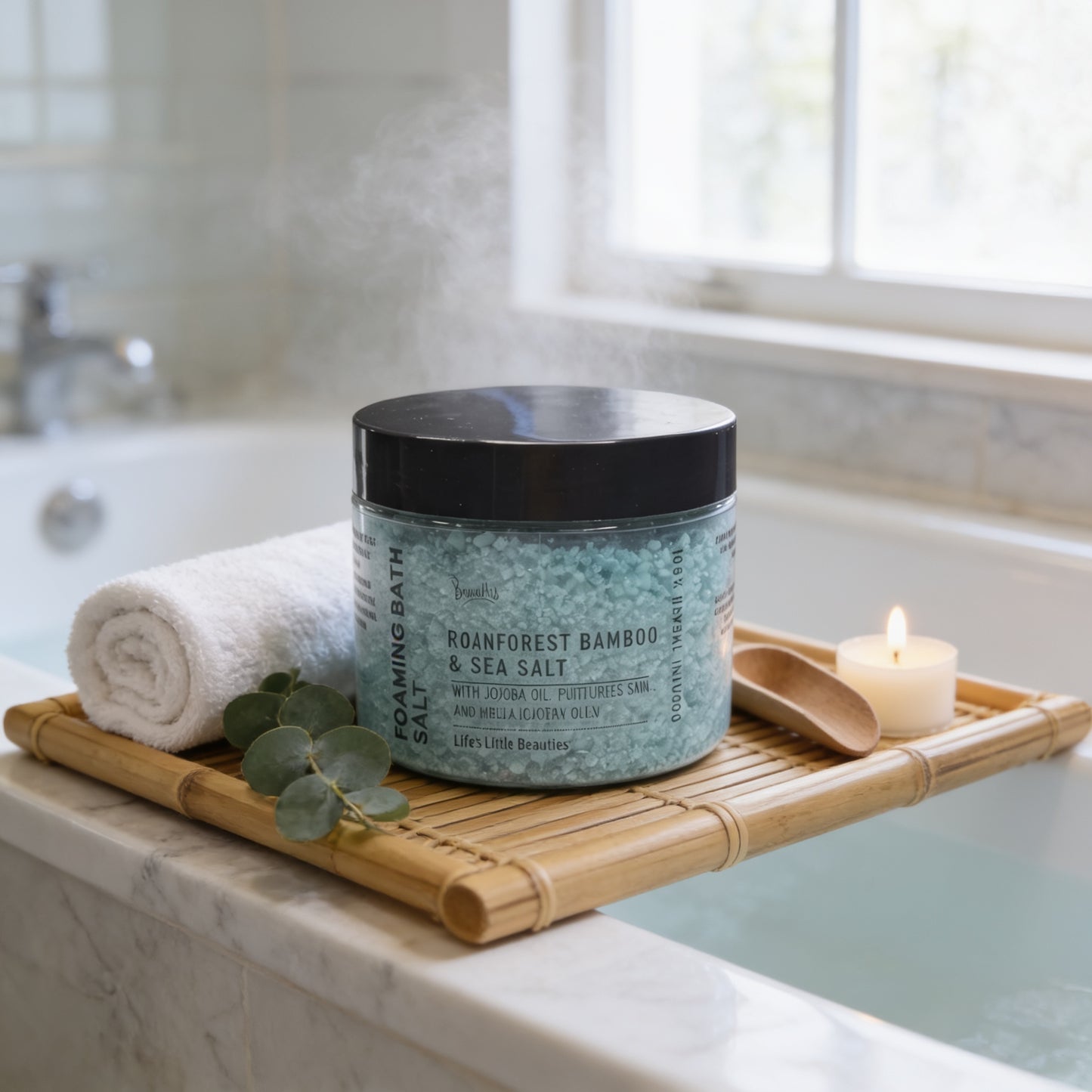 A jar of Roanforest Bamboo & Sea Salt Soaking Bath Salt is placed on a bamboo tray, with a lit candle and a rolled white towel nearby, in a bathroom setting.