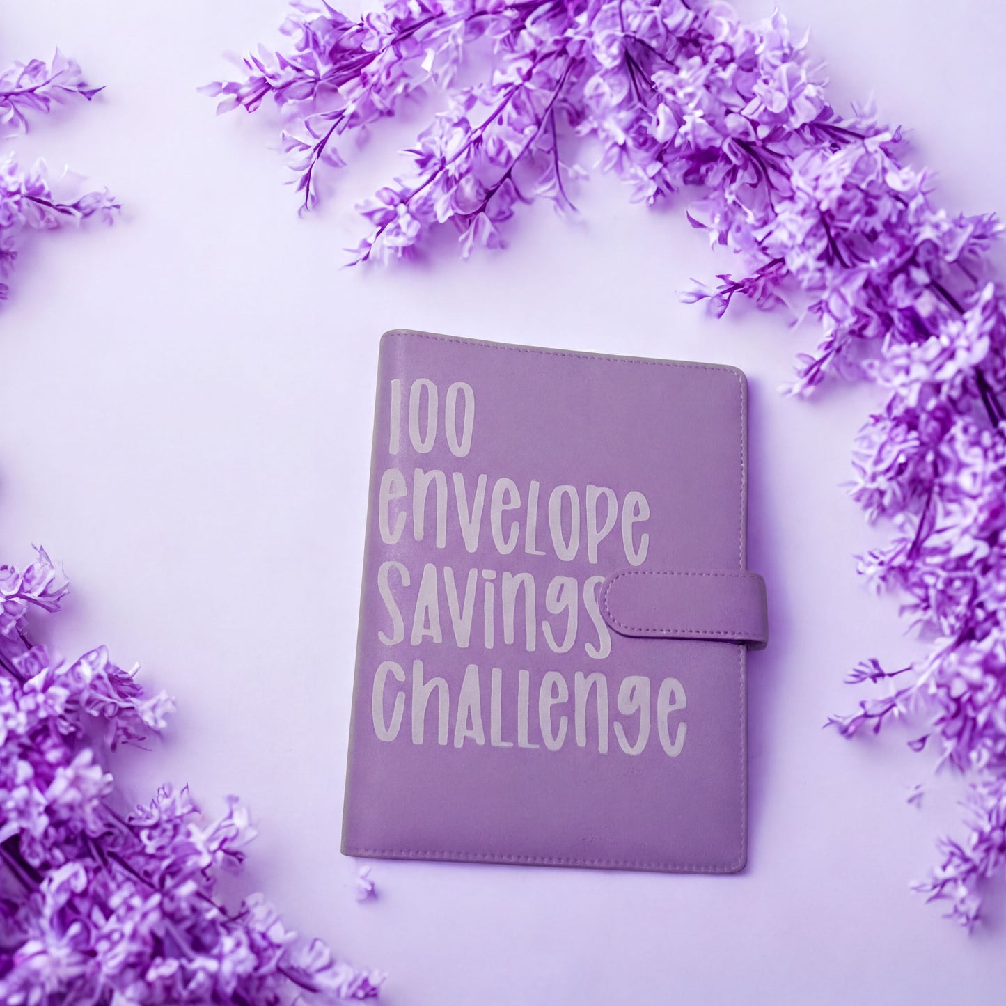 A purple notebook with the text "100 envelope savings challenge" printed on it, surrounded by purple flowers on a light purple background.