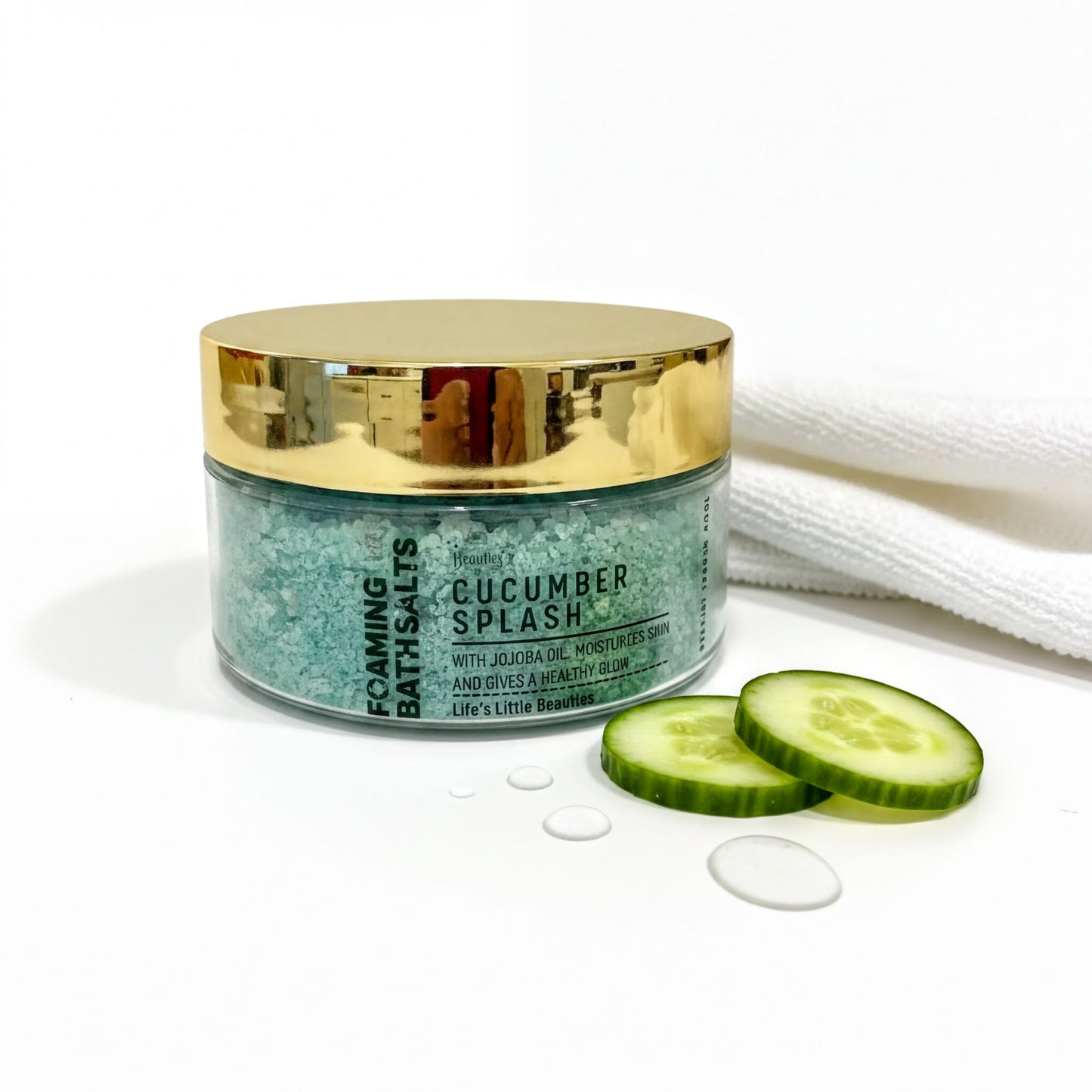 A jar of cucumber splash bath salt with a gold lid, accompanied by sliced cucumbers and a white towel on a white surface.
