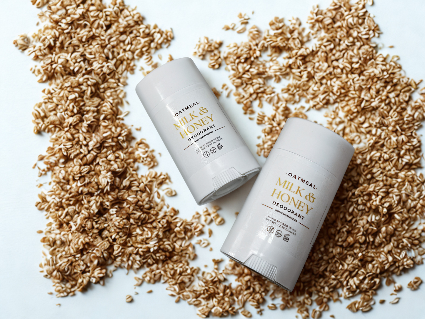 Two white deodorant sticks with the text "OATMEAL MILK & HONEY DEODORANT" are placed on a white surface, surrounded by scattered oat flakes.
