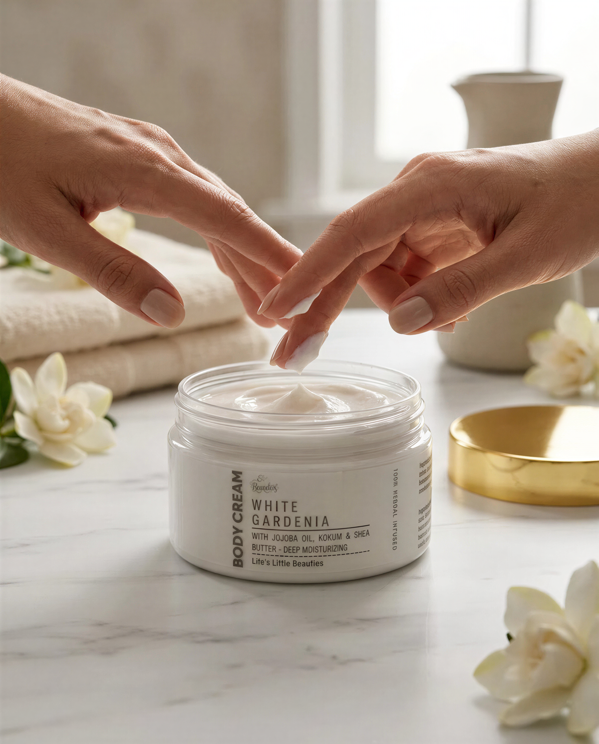 A person's hands applying a white body cream to their hands, with a jar of the cream in the foreground and white flowers in the background.