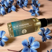 A bottle of "Life's Little Beauty" Blue Hawaiian Post-Shower Spray" surrounded by blue flowers on a wooden surface.