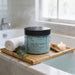 A jar of Roanforest Bamboo & Sea Salt Soaking Bath Salt is placed on a bamboo tray, with a lit candle and a rolled white towel nearby, in a bathroom setting.