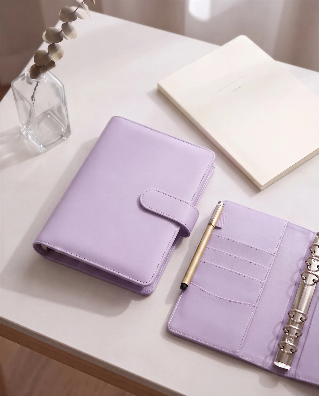 a purple leather-bound notebook, a pen, and a small vase with dried flowers on a white surface.