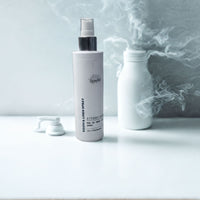 a bottle of "Room & Linen Spray" with a white cap, placed on a white surface against a backdrop of a white wall with smoke or steam coming from it.