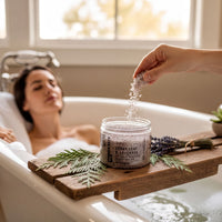 A person relaxing in a bathtub, surrounded by a wooden tray with a jar of bath salt, a candle, and some greenery.
