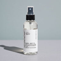A bottle of "Dark Arctic" body spray with a black cap and a clear pump bottle, standing on a gray surface against a light blue background.