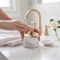 A person's hands are shown applying a white cream to their face, with a jar of the cream and a small bowl of the cream in the background.