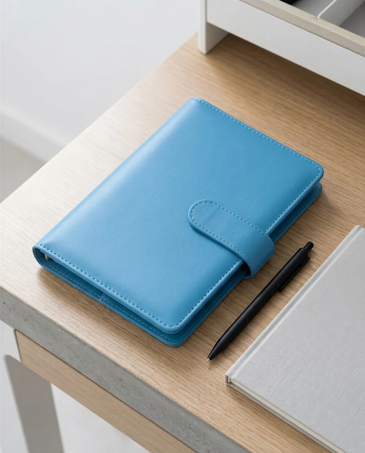 A blue leather-bound notebook and a black pen are placed on a wooden desk, with a gray notebook and a white drawer visible in the background.