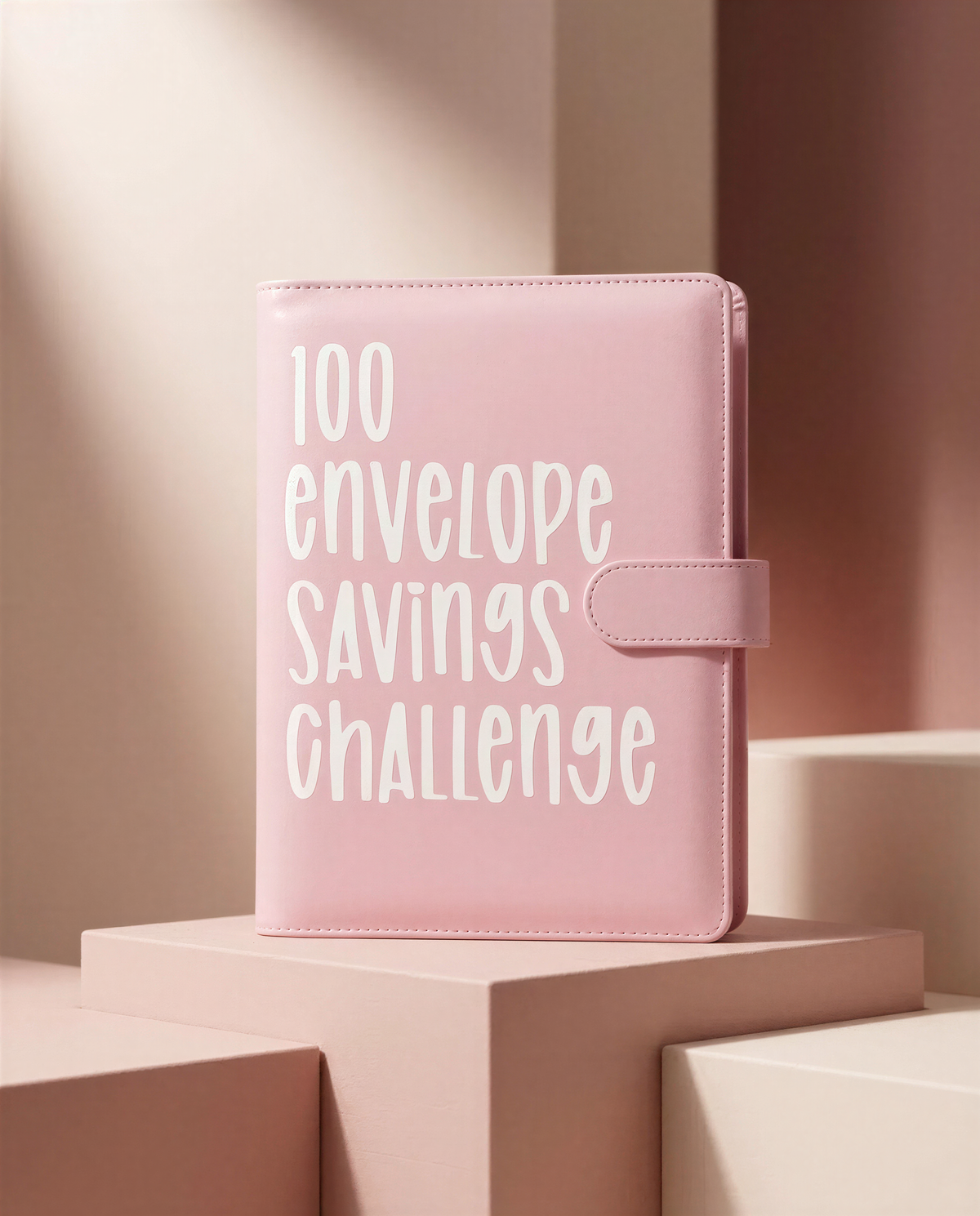 A pink leather-bound notebook with the text "100 envelope savings challenge" printed on it, placed on a pink platform against a pink background.