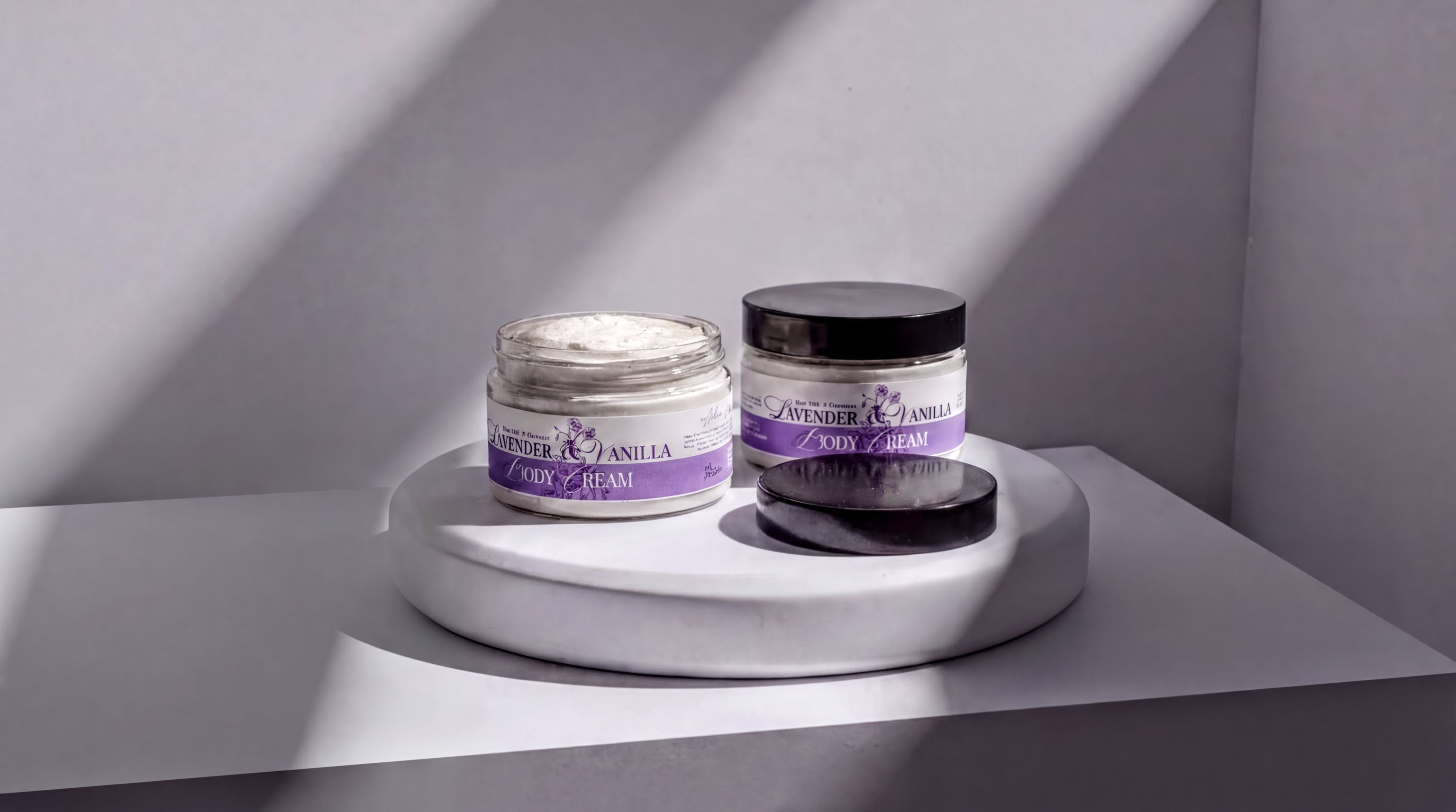 Two jars of lavender and vanilla body cream are placed on a white pedestal against a white wall, with sunlight casting shadows on the pedestal.