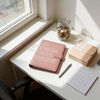 a pink notebook with the text "100 envelope savings challenge" on it, placed on a white desk next to a jar filled with coins, a pen, and a calendar.