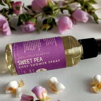 a bottle of "Sweet Pea Post-Shower Spray" surrounded by pink roses and petals on a white surface.