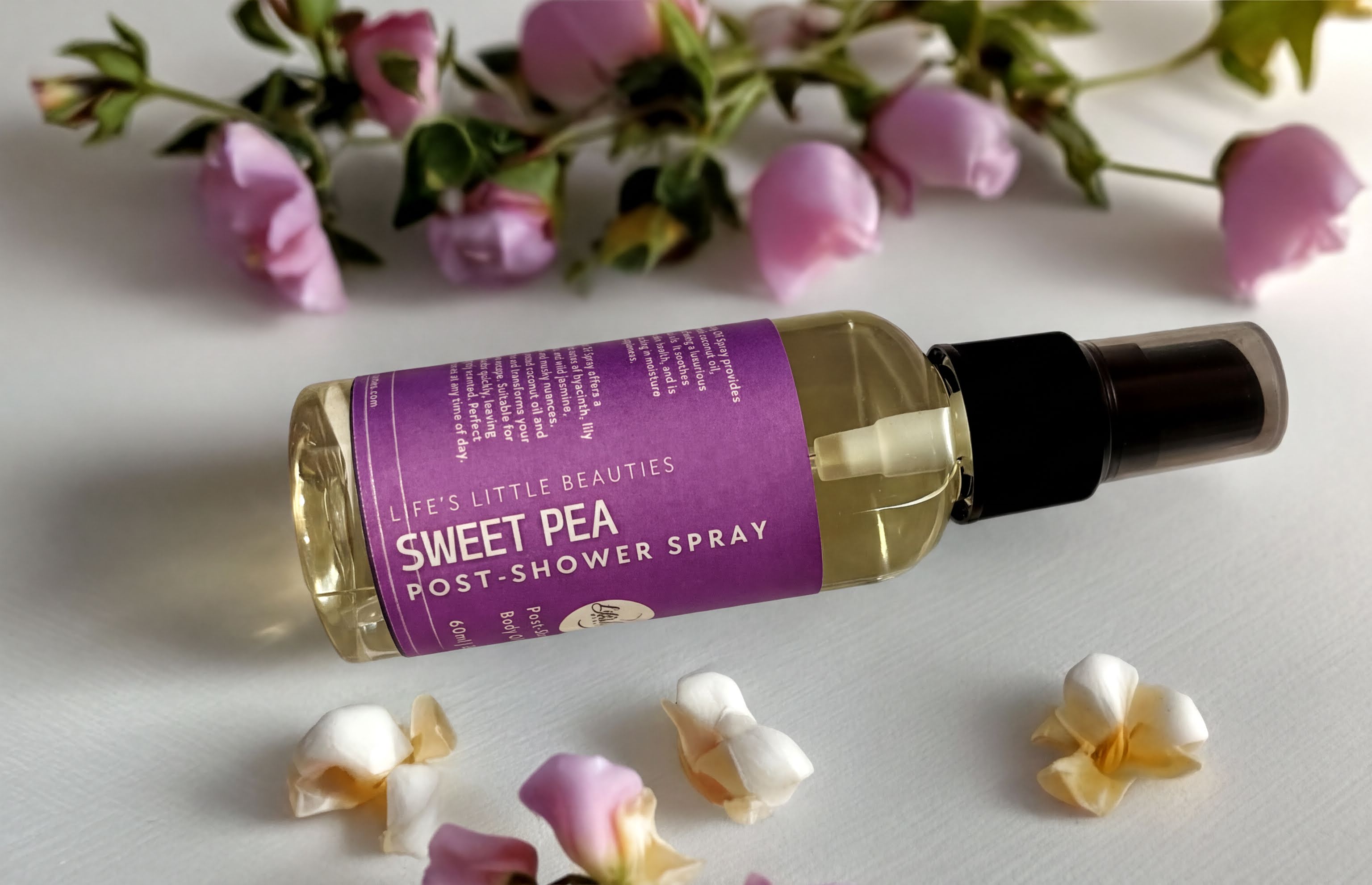 a bottle of "Sweet Pea Post-Shower Spray" surrounded by pink roses and petals on a white surface.