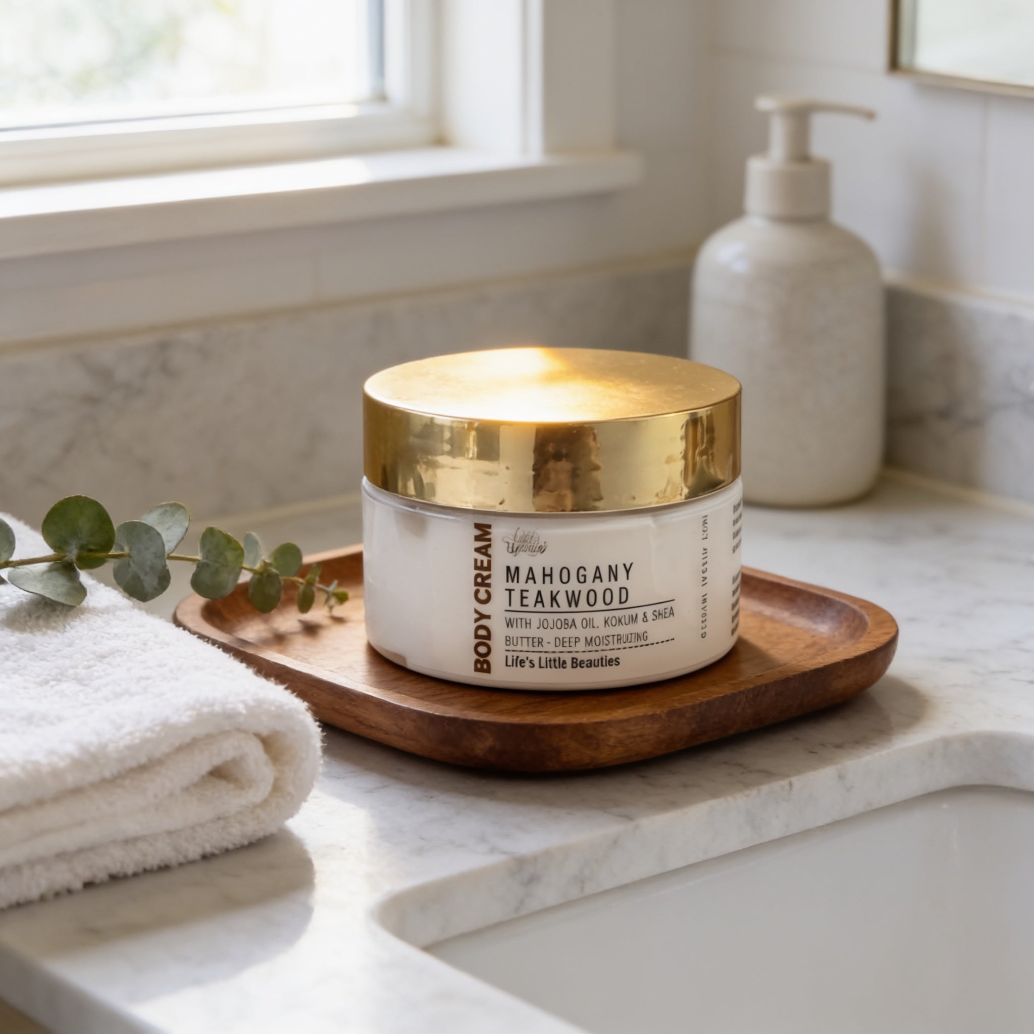 A jar of Mahogany Teakwood Body Cream with Jojoba Oil sits on a wooden tray atop a white marble countertop, accompanied by a white pump bottle and a green plant.