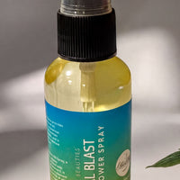 A bottle of "Tropical Blast" body spray is shown on a white surface, with a plant leaf and a piece of fruit visible in the background.