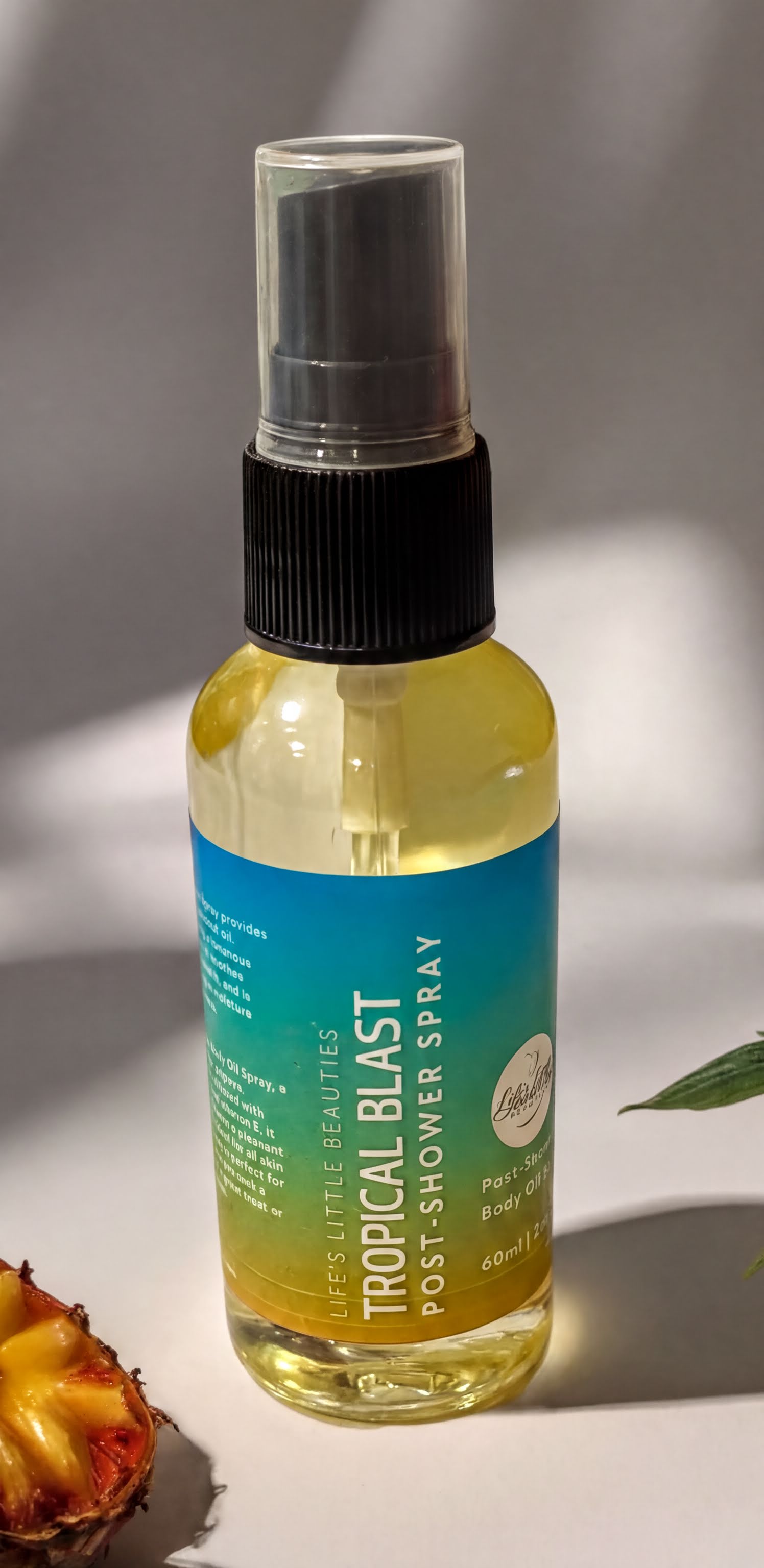 A bottle of "Tropical Blast" body spray is shown on a white surface, with a plant leaf and a piece of fruit visible in the background.