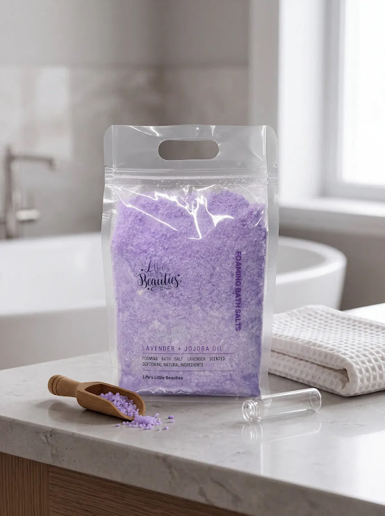 A clear plastic bag filled with lavender-colored bath salt is placed on a bathroom counter, with a wooden scoop and a glass bottle of the same bath salt nearby.