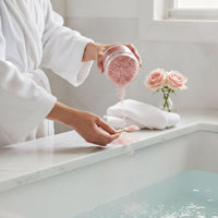 A person in a white robe is pouring pink bath salt into a bathtub, with a vase of pink roses nearby.