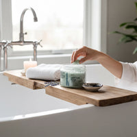 A person is sitting in a bathtub, surrounded by various items, including a jar of bath product, a candle, and a wooden tray.