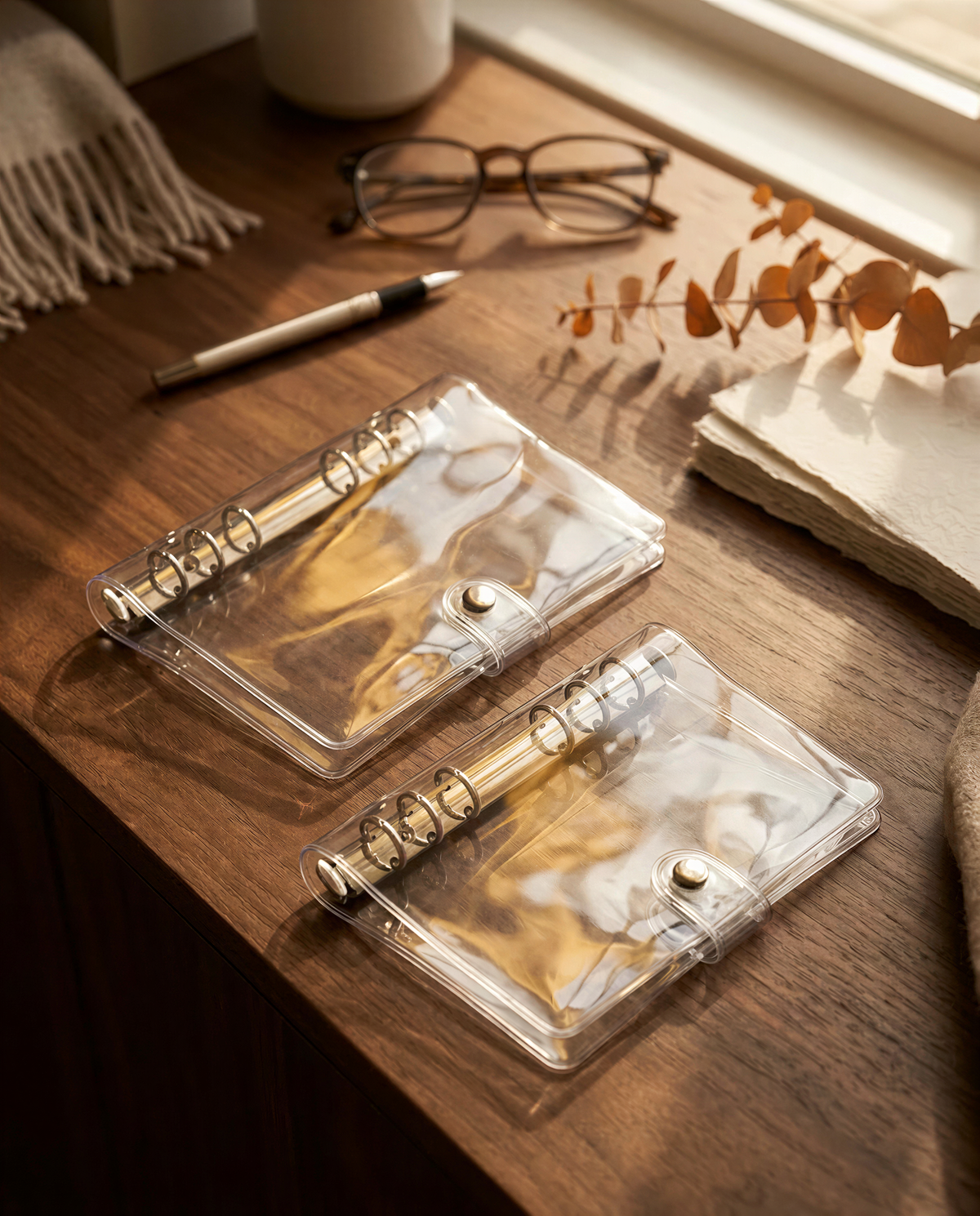 a wooden desk with various items on it, including two transparent planners, a pair of glasses, and some dried flowers.