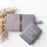 A purple leather-like organizer with metal rings and a cover that says "100 envelope savings challenge" is placed on a white surface next to a pile of beige fuzzy material.
