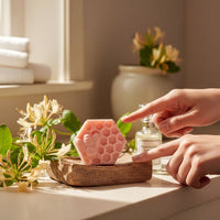 A person's hands are pointing at a pink soap bar with a honeycomb design, surrounded by white flowers and a wooden tray on a countertop.