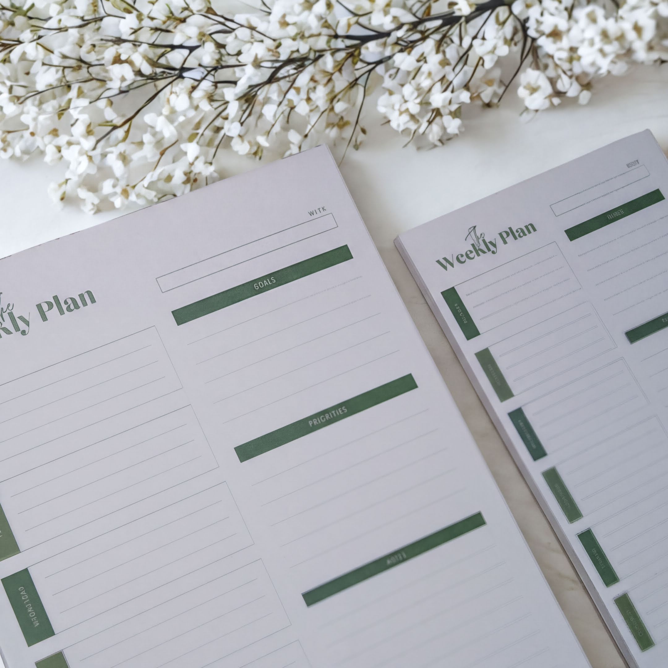 two planners or notebooks with a white background and green lines, placed on a white surface. There are also some white flowers in the background.