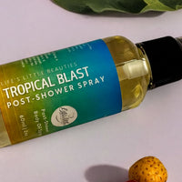 A bottle of "Life's Little Beauties Tropical Blast Post-Shower Spray" is shown on a white surface, surrounded by a green leaf and a small fruit.