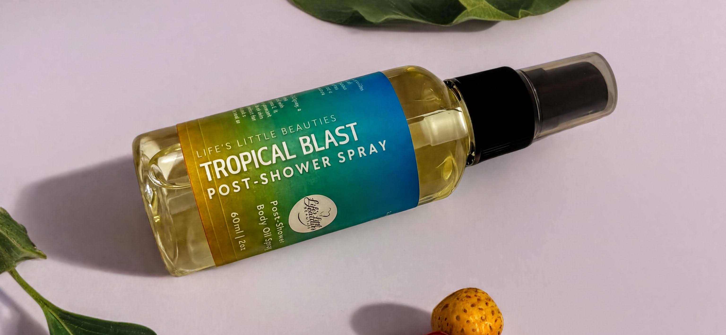 A bottle of "Life's Little Beauties Tropical Blast Post-Shower Spray" is shown on a white surface, surrounded by a green leaf and a small fruit.