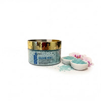 a jar of Ocean Mist bath salt with a gold lid, surrounded by small white bowls filled with the same blue-green bath salt, and a few pink orchid petals.