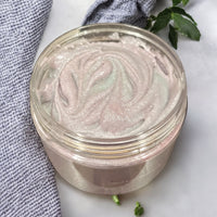 A clear jar filled with a pink, creamy substance, possibly a homemade body scrub or lotion, sits on a white surface next to a blue and white striped cloth and a small green plant.