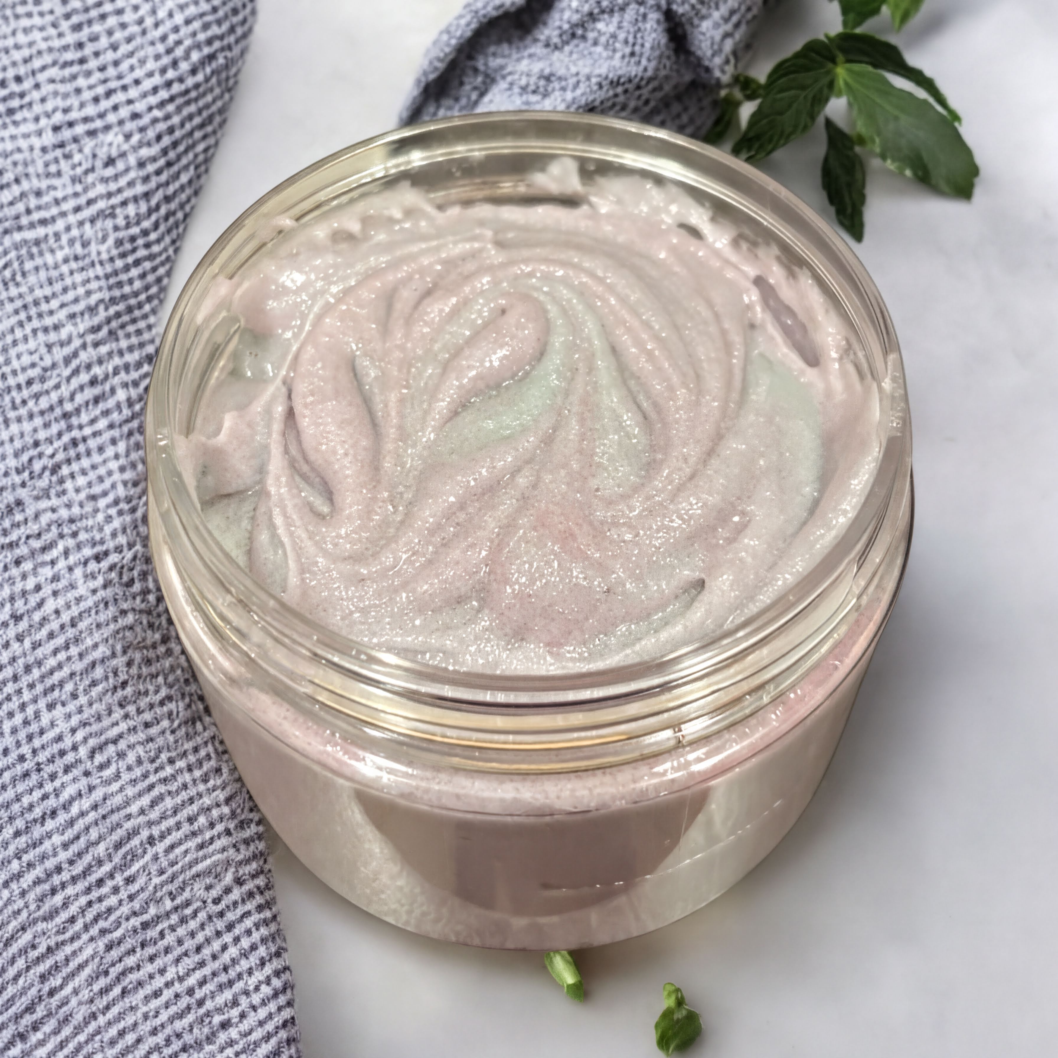 A clear jar filled with a pink, creamy substance, possibly a homemade body scrub or lotion, sits on a white surface next to a blue and white striped cloth and a small green plant.