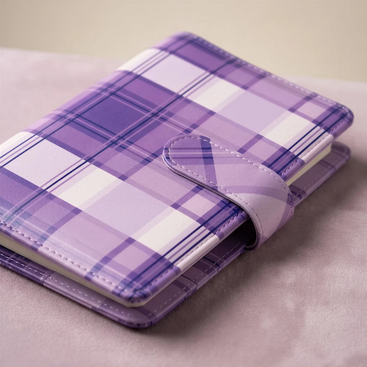 A purple and white plaid notebook with a leather-like texture and a leather-like clasp, resting on a beige surface.