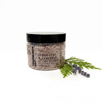 A jar of Cedar Leaf & Lavender Soaking Bath Salt, surrounded by a sprig of lavender and a fern leaf, against a white background.