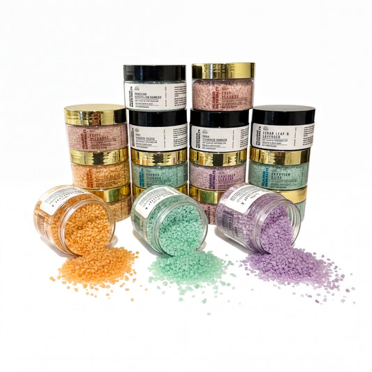 a collection of jars and containers filled with various colored bath salt crystals, including orange, green, purple, and lavender, spilling out onto a white background.