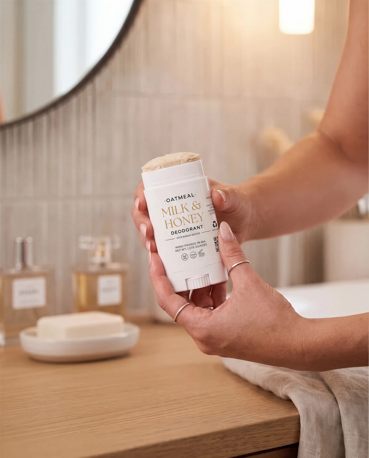 A person's hands holding a white and gold OATMEAL MILK & HONEY DEodorant stick in a bathroom setting.
