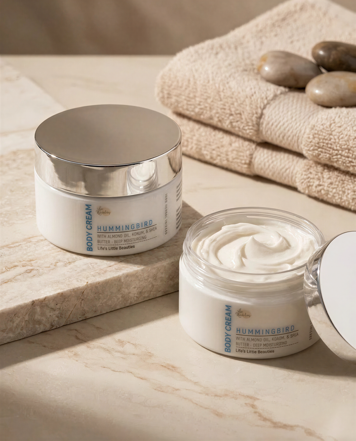 two jars of body cream, one with a lid and the other with the lid off, placed on a marble surface. There are also some rocks and a towel in the background.