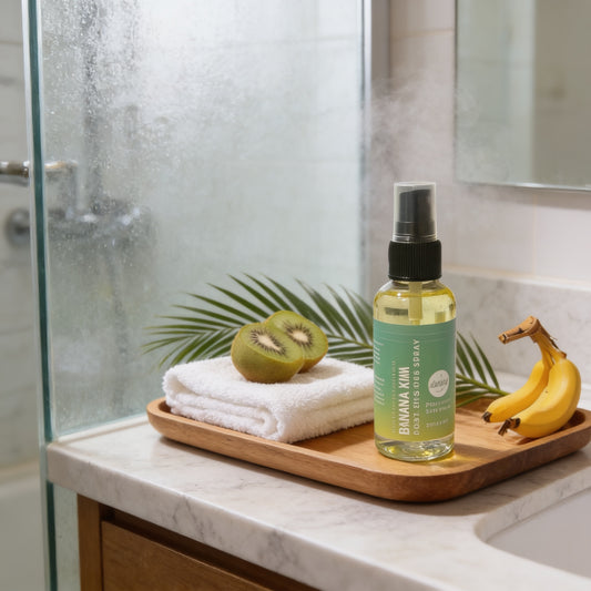 A bathroom counter with a bottle of Banana & Kiwi Body Mist, a folded towel, a banana, and a leafy green plant.