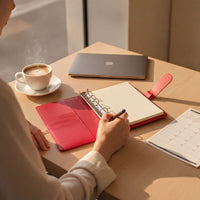 A person is sitting at a desk, writing in a red notebook with a pen, while a laptop and a cup of coffee are nearby.