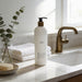 A bottle of body wash sits on a marble countertop next to a sink, with a vase of greenery and a folded towel nearby.