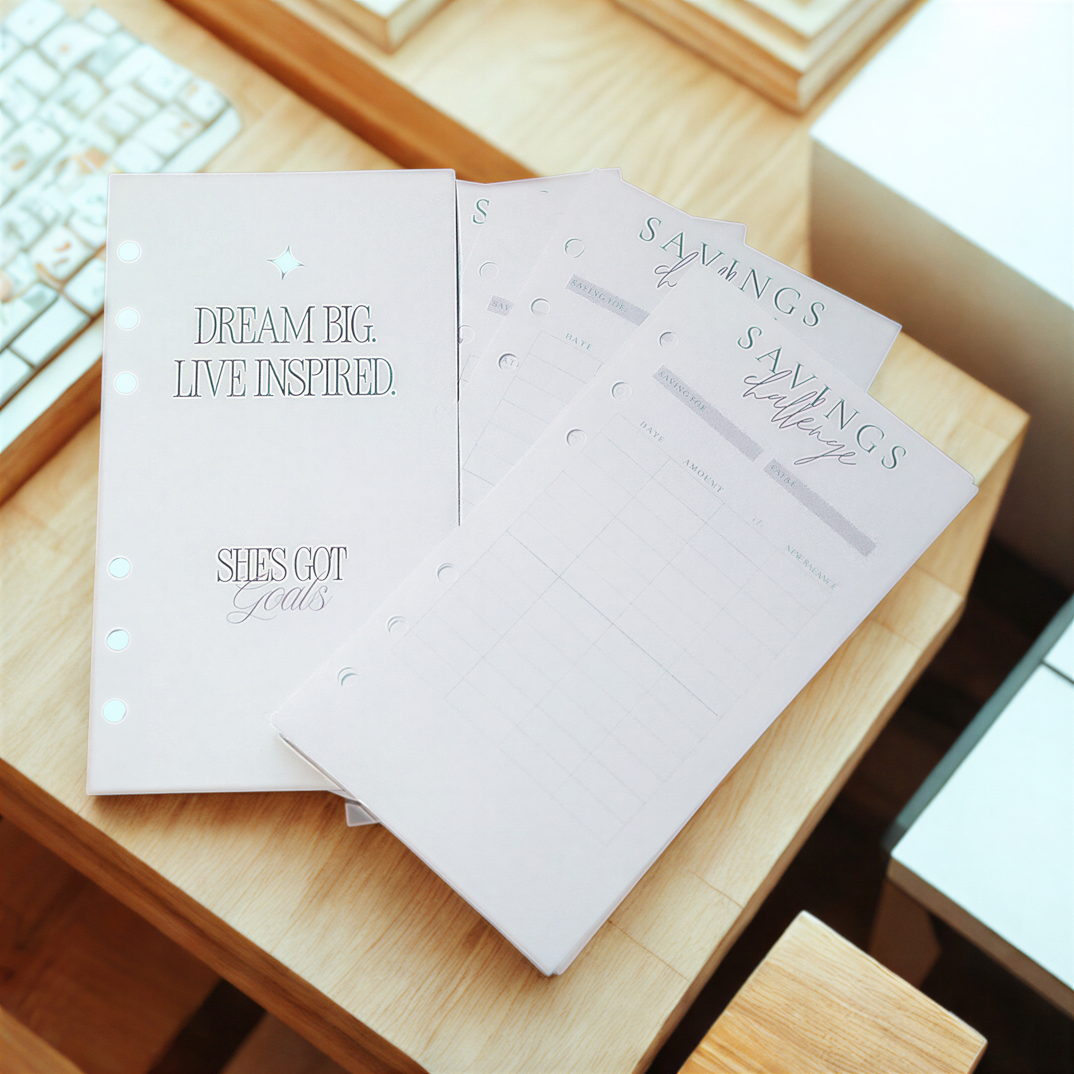 a desk with a notebook and a stack of blank planners or notebooks. The notebook has a title that reads "DREAM BIG LIVE INSPIRED" and "SHES GOT GOALS". The planners or notebooks have a title that reads "SAVINGS" and "Saving Planner".