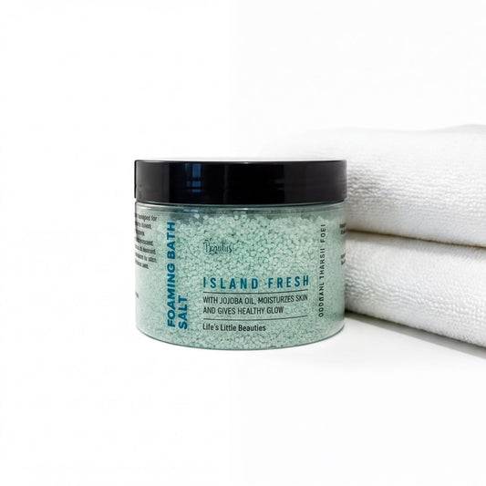 A jar of "Island Fresh" bath salt with jojoba oil, moisturizes skin, and gives healthy glow, placed on a white surface next to a folded white towel.