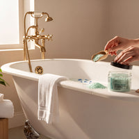A person's hands are pouring a green bath bomb into a white bathtub filled with water, surrounded by various bath products and decorative elements.