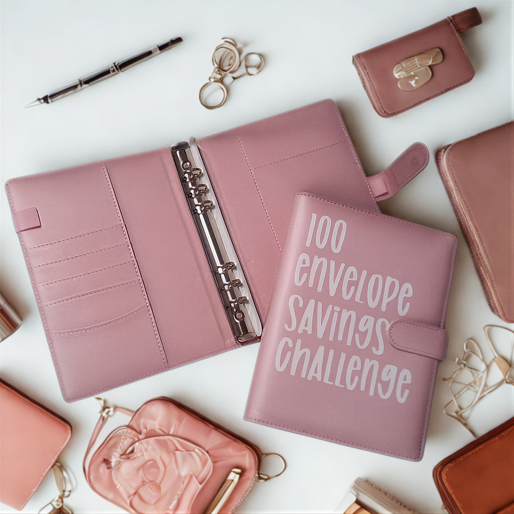Pink 100 envelope savings challenge planner with matching wallet and accessories on white desk
