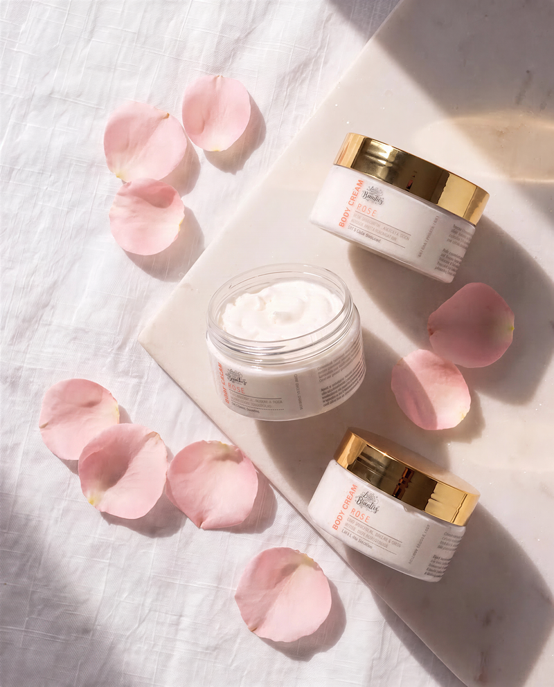 three jars of body cream with gold lids, along with a few pink rose petals scattered around them.