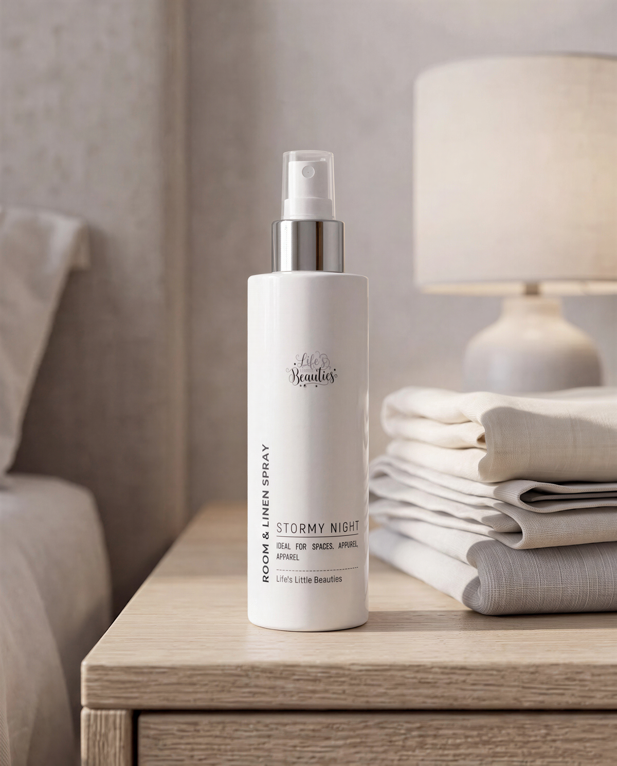 A bottle of "Stormy Night" linen spray is placed on a wooden nightstand, with a stack of folded white linens and a lamp in the background.