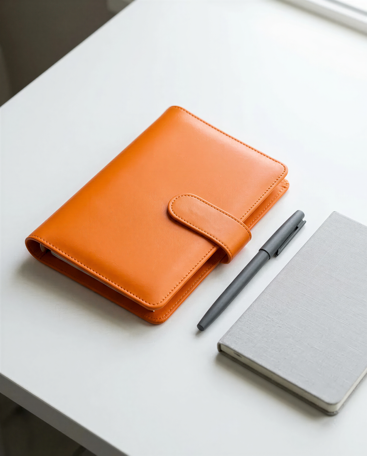 An orange leather-bound notebook, a black pen, and a gray notebook are arranged on a white surface.