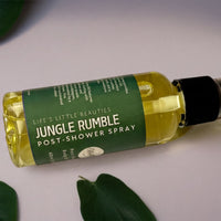 A bottle of Jungle Rumble Post-Shower Spray surrounded by green leaves on a white background.